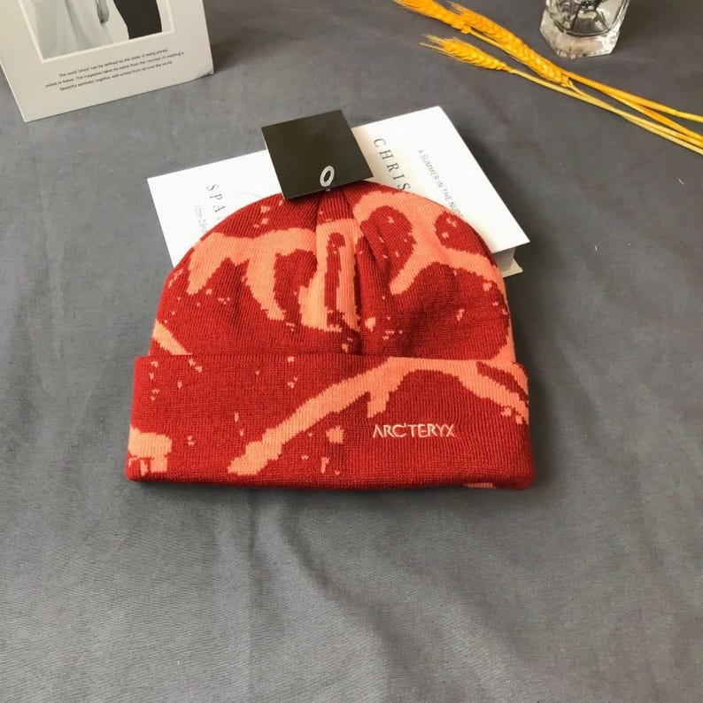 Peut inclure: Bonnet rouge et orange avec un motif unique et le nom de la marque "Arc'teryx" brod&eacute; sur le devant. Le bonnet est pli&eacute; au niveau du bord et une &eacute;tiquette est attach&eacute;e. Le bonnet est pos&eacute; sur un livre.