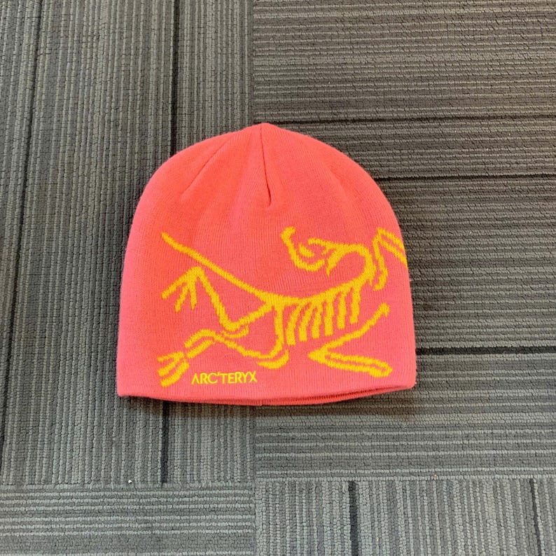 May include: A coral-colored beanie with a yellow graphic of a stylized creature and the text "ARC'TERYX" at the bottom. The hat is made of a soft, knitted material and is designed to keep the head warm.