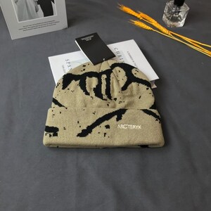 Peut inclure: Un bonnet beige avec des motifs noirs abstraits et la marque "Arc'teryx" brod&eacute;e en blanc. Le chapeau a un bord repli&eacute; et une &eacute;tiquette attach&eacute;e. Le bonnet est pos&eacute; sur un livre blanc et une surface grise.