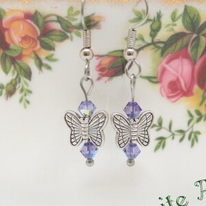 May include: Silver butterfly earrings with purple tanzanite AB crystals hanging from a white teacup.