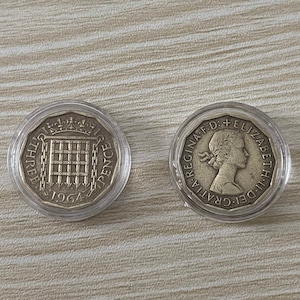May include: Two silver British coins in plastic cases. The coin on the left is a three pence coin with the year 1964 and a coat of arms. The coin on the right is a silver coin with the profile of Queen Elizabeth II and the text "GRATIA DEI. D.G. REGINA. F.D. ELIZABETH II."