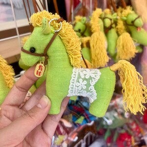 Può includere: Un cavallo fatto a mano in tessuto verde con criniera e coda gialle. Il cavallo ha una briglia marrone e una coperta da sella in pizzo bianco. Un piccolo cartellino color oro con caratteri cinesi pende dalla briglia. Il cavallo è un portachiavi.