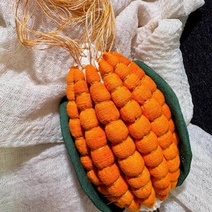 Puede incluir: Un objeto decorativo hecho a mano con forma de mazorca de maíz. El maíz está hecho de hilo naranja, con una cáscara de tela verde. En la parte superior hay hilos dorados y beige. Se coloca sobre una tela blanca texturizada.