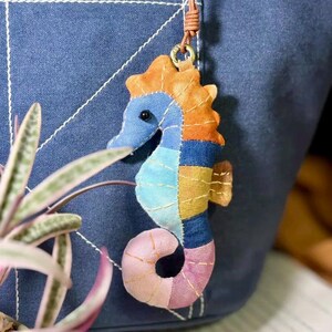 May include: A colorful, handmade seahorse charm. The seahorse is crafted from fabric scraps in shades of blue, orange, yellow, and pink, stitched together. It hangs from a brown leather cord, attached to a metal ring.