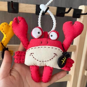 Può includere: Ornamento a forma di granchio rosso fatto a mano con dettagli bianchi e un volto sorridente. Il granchio ha grandi chele rosse e una piccola tasca nera. È attaccato a un filo bianco per essere appeso.