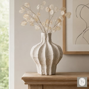 May include: A textured, off-white ceramic vase with a distinctive, ribbed design. The vase contains delicate, dried seed pods in a light beige hue. The vase is placed on a wooden surface, with framed artwork visible in the background.