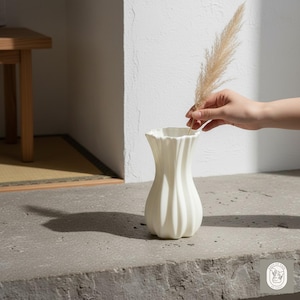 Puede incluir: Un jarrón de cerámica blanco con un diseño acanalado. Una mano coloca un tallo de hierba ornamental seca en el jarrón. El jarrón está sobre una superficie de piedra gris. El jarrón tiene una base redondeada y una parte superior con volantes.