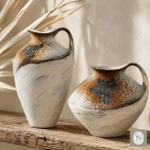 May include: Two ceramic vases with a rustic, handcrafted appearance. The vases are off-white with brown and black accents, featuring a unique handle design. The taller vase is approximately 12 inches tall, while the shorter one is about 8 inches.