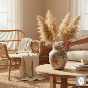 May include: A beige ceramic vase with a textured finish holds several dried pampas grass stems. The vase sits on a round wooden coffee table. A wicker chair and a beige blanket are in the background.