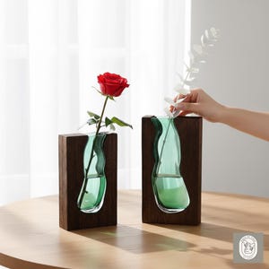 Könnte beinhalten: Zwei dekorative Vasen mit Holzrahmen und einem teal-farbenen Glaseinsatz. Eine Vase enthält eine rote Rose, die andere wird mit weißen Blumen gefüllt. Die Vasen stehen auf einem Holztisch.