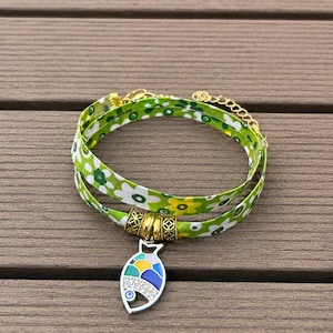 Pode incluir: Uma pulseira de enrolar com uma fita de tecido verde com um padrão floral branco e amarelo. A pulseira inclui três contas de metal dourado e um pingente em forma de peixe prateado com segmentos de esmalte colorido e pequenos cristais.