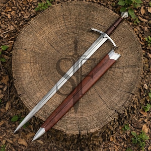 Puede incluir: Una espada de estilo medieval con una hoja plateada y un mango envuelto en cuero marrón. La espada descansa sobre un tocón de árbol de madera junto a una vaina de cuero marrón con detalles plateados. Es una réplica histórica.