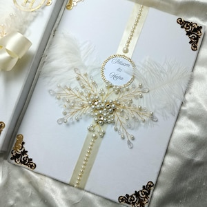May include: White wedding invitation box with gold filigree corners and a pearl and crystal embellishment. A cream ribbon and a string of pearls adorn the box. The box has a circular tag with the names "Ibtisam & Hafsa".