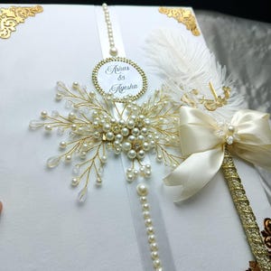 May include: A white rectangular box featuring gold corner accents, a central vertical white ribbon adorned with a string of pearls, and a circular tag with the text "Ashas & Ayesha". A gold wire spray with white pearls and a cream satin bow with a white feather.