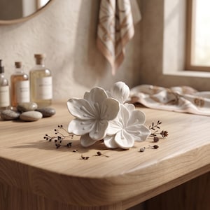 May include: A white ceramic floral sculpture with detailed petals and a bud, resting on a light wood surface. Decorative sprigs and small stones are scattered around the sculpture. The background includes bottles and a towel.