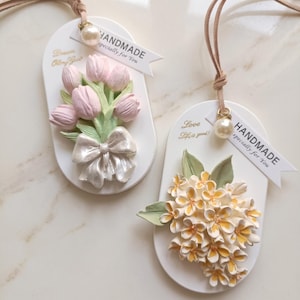 May include: Two white, oval-shaped handmade ornaments with floral designs. One features pink tulips with a bow, the other has yellow and white flowers. Each has a ribbon, a pearl, and the words "HANDMADE specially for You".