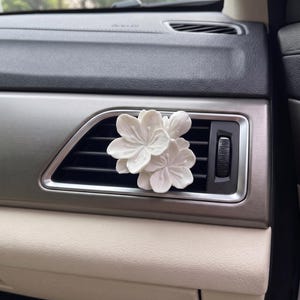 Puede incluir: Un ambientador floral blanco para coche está enganchado a una rejilla de ventilación del coche. El ambientador está hecho de múltiples flores blancas. El interior del coche presenta una rejilla de ventilación plateada, un salpicadero negro y un panel inferior beige.