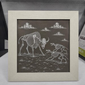 May include: A square framed artwork featuring a white line drawing on a dark grey background. The illustration depicts the skeletal remains of a large bison facing two smaller canine skeletons in a field under several stylised white clouds.