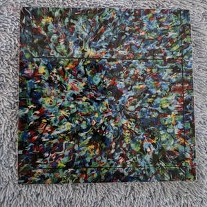 May include: Square coaster with an abstract design. The coaster features a colorful pattern of blue, green, red, and yellow brushstrokes. The edges are outlined with a thin black border.