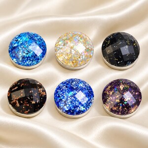 Op de afbeelding: Zes ronde, gefacetteerde cabochons gerangschikt in twee rijen. De bovenste rij bevat een felblauw, een goud-wit en een zwart exemplaar. De onderste rij bevat een donkerbruin, een felblauw en een dieppaars exemplaar, allemaal met metallic spikkels.