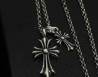 Puede incluir: Un collar de metal plateado con una cadena de eslabones redondos y dos colgantes en forma de cruz. La cruz más grande tiene un acabado pulido con extremos ensanchados, mientras que la cruz más pequeña está unida a la misma argolla.