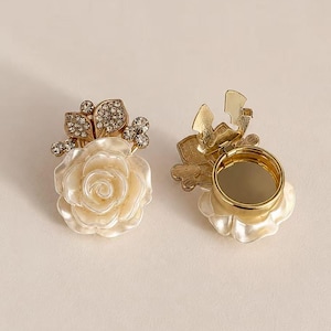 May include: Two gold-toned metal ring accessories featuring a cream-colored rose design. One ring shows the front with a floral motif and clear crystal accents on the leaves, while the other shows the underside with a circular adjustable band.
