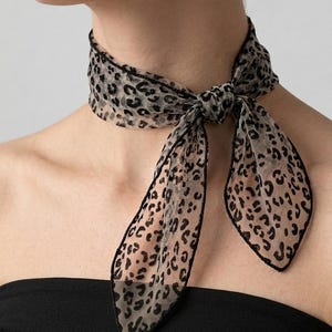 May include: A sheer scarf with a leopard print pattern. The scarf is tied around the neck in a bow, with the ends hanging down. The edges of the scarf are trimmed in black.