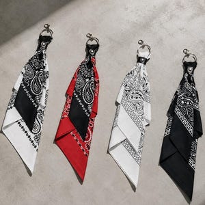 Könnte beinhalten: Vier Schlüsselanhänger mit gefalteten Stoffbandanas mit Paisleymustern in Weiß, Rot und Schwarz. Jedes Bandana ist mit einer schwarzen, lederartigen Lasche, die mit einer silbernen Niete befestigt ist, an einem silberfarbenen Ring und Karabiner befestigt.