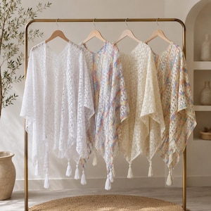 Könnte beinhalten: Vier leichte Ponchos mit V-Ausschnitt hängen an einem goldfarbenen Metallständer. Der erste ist reinweiß, der zweite hat ein mehrfarbiges Muster, der dritte ist cremefarben und der vierte hat ein Blumenmuster. Jedes Kleidungsstück hat eine Spitzenstruktur.
