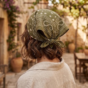 Könnte beinhalten: Olivgrünes Bandana mit weißem Blumen- und Paisley-Muster, das am Hinterkopf gebunden ist. Das Bandana ist aus einem weichen Stoff gefertigt und wird über kurzem, welligem braunem Haar getragen. Die Person trägt ein hellbeige Leinenhemd.