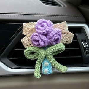 Könnte beinhalten: Ein handgefertigter Häkel-Autolüftungsclip mit drei lila Rosen, einer cremefarbenen Spitzen-Schleife und einer grünen Schleife mit hellblauem Akzent und zwei Perlen. Das florale Design ist an einer Autolüftung befestigt.