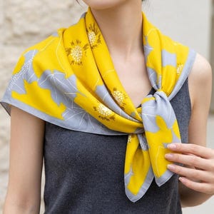 Könnte beinhalten: Ein quadratisches Tuch mit leuchtend gelbem Hintergrund und einem sich wiederholenden Muster aus stilisierten grauen Blättern und weißen kreisförmigen Blumenmotiven. Das Tuch ist vorne geknotet und hat einen grauen Rand am Stoffsaum.