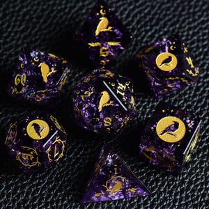 Könnte beinhalten: Ein Set aus sieben polyedrischen Würfeln aus durchscheinendem violettem Harz mit eingearbeitetem Glitzer. Jeder Würfel zeigt goldfarbene Gravuren von Raben, Mondsicheln, Sternen und Zweigen. Die Zahlen sind in einer stilisierten Serifenschrift gehalten.