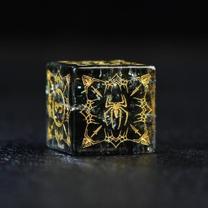 May include: A translucent black six-sided die featuring intricate gold-colored engravings on each face. The visible side displays a detailed spider icon centered within a decorative frame of pointed shapes and lines.