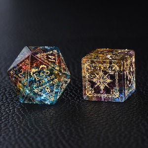 May include: Two decorative dice. One is a multi-faceted, multi-colored die with gold accents and the text "E&S 07.18.2026". The other is a cube-shaped die with gold starburst and geometric designs.