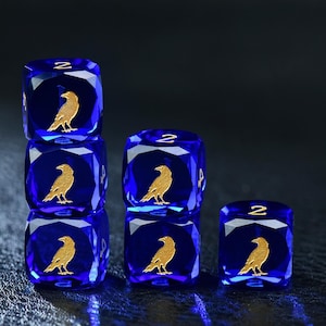 Könnte beinhalten: Ein Set aus fünf durchscheinenden, tiefblauen sechsseitigen Würfeln mit facettierten Kanten. Jeder Würfel zeigt eine goldfarbene Rabensilhouette auf der Vorderseite und die eingravierte goldene Zahl zwei auf der Oberseite. Die Würfel sind gestapelt.