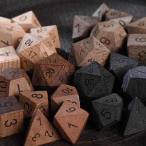 Op de afbeelding: Een verzameling houten polyhedrale dobbelstenen in verschillende tinten bruin en grijs. De dobbelstenen hebben verschillende vormen, waaronder D4, D6, D8, D10, D12, D20 en D100. De cijfers zijn in een donkere kleur geëtst.