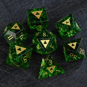 May include: Set of seven green and gold polyhedral dice. Each die features a gold triangle symbol and gold numbering. The dice are made of a translucent green material with a marbled effect, and are designed for tabletop gaming.