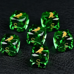 Pode incluir: Um conjunto de seis dados de seis faces em verde esmeralda translúcido. Cada dado apresenta um ícone de corvo na cor dourada na face superior e o número cinco numa fonte dourada na face frontal. Os dados têm arestas facetadas e um acabamento polido.