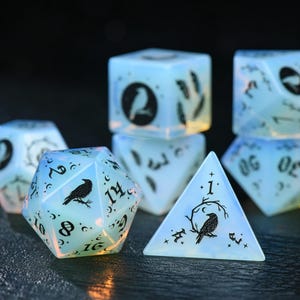 May include: A set of polyhedral dice made of a translucent, milky blue material. Each die features black ink engravings of ravens, crescent moons, stars, and feathers. The numbers are displayed in a decorative serif font across the various faces.