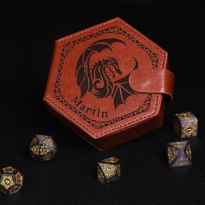 Puede incluir: Una caja de dados de cuero marrón con un diseño de dragón y el nombre "Martin" grabado. Varios dados de múltiples caras con detalles dorados están dispersos alrededor de la caja, para juegos de mesa.