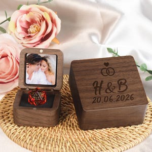 May include: A dark wood ring box with a hinged lid featuring a photograph of a couple inside. The exterior lid is engraved with two interlocking rings, a heart, the letters H & B, and the date 20.06.2026. A red translucent 20-sided die sits inside.