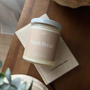 May include: A glass jar candle with a light grey lid and a beige label that reads "Fresh Bread." The candle is 213 g and made with coconut soy. It sits on a small, beige book with the text "A FIVE-YEAR MEMORY BOOK."