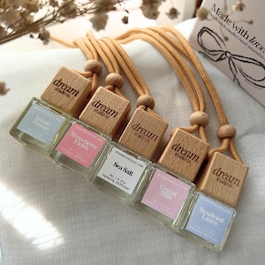 May include: A selection of hanging car air fresheners with wooden lids and beige cords. The glass bottles display labels with scent names such as "Island Breeze," "Strawberry Fields," and "Sea Salt." The wooden lids are branded with "dream studio co."