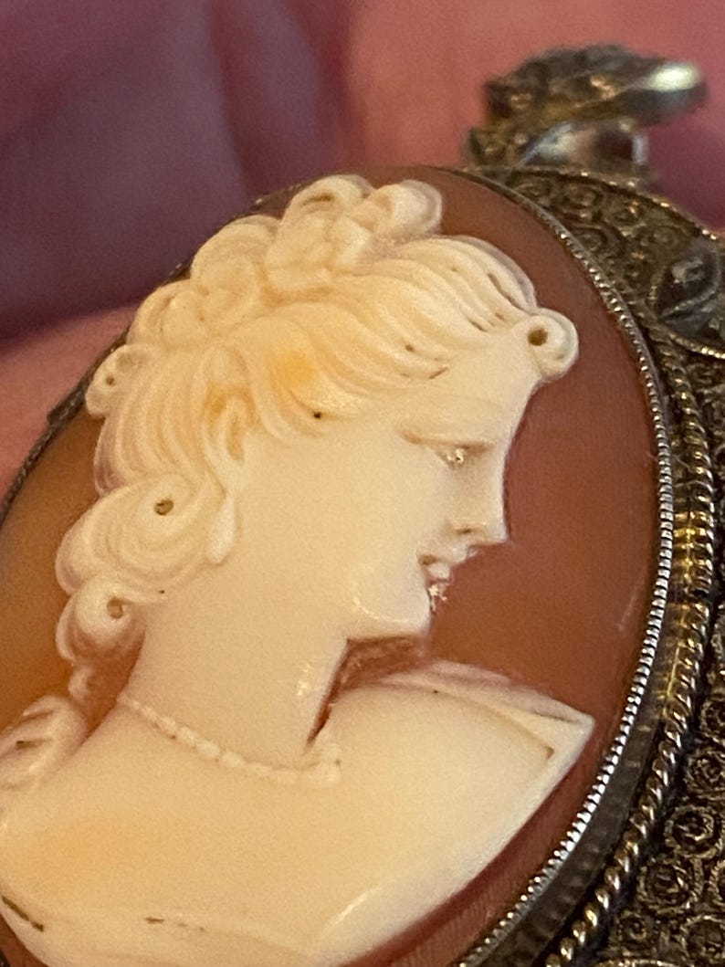 May include: Close-up of a cameo pendant featuring a profile of a woman with detailed hair and a delicate necklace. The cameo is set in a silver filigree frame, showcasing the intricate design and craftsmanship of the jewelry.