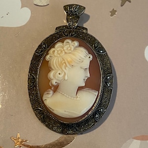 May include: Oval cameo pendant featuring a profile of a woman with a pearl necklace and ornate hairstyle. The cameo is set in a detailed, antique-style metal frame with a decorative bail. The background is a warm, reddish-brown color.