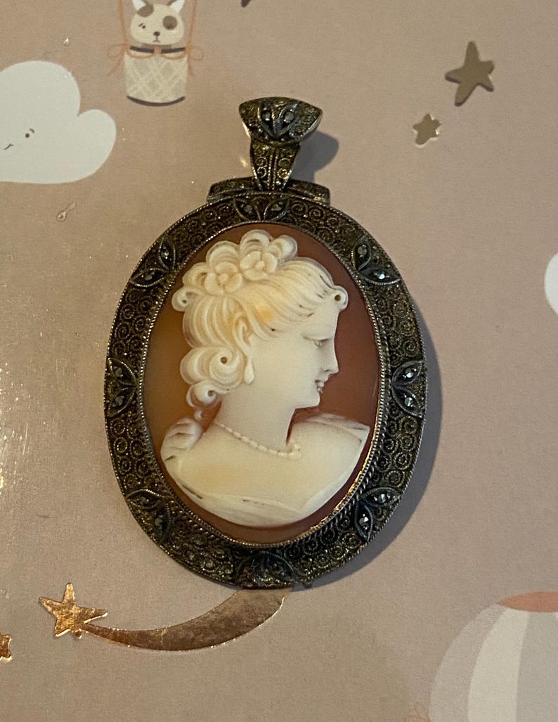 May include: Oval cameo pendant featuring a profile of a woman with a pearl necklace and ornate hairstyle. The cameo is set in a detailed, antique-style metal frame with a decorative bail. The background is a warm, reddish-brown color.