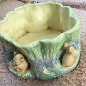 Könnte beinhalten: Ein Keramik-Pflanzgefäß mit hellgrüner und blauer Außenseite, das zwei kleine Hasenfiguren zeigt. Der Innenraum ist hellgelb. Der Pflanztopf hat eine einzigartige, organische Form, die ein natürliches Thema suggeriert.