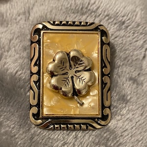 May include: A rectangular brooch with a gold-colored frame and a four-leaf clover design. The clover is silver-colored and set against a cream-colored background. The frame has an ornate, scrolling design.
