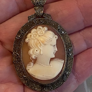 May include: Oval cameo pendant featuring a profile of a woman with a floral hairpiece. The cameo is set in a decorative silver-toned frame with marcasite accents. The pendant has a detailed bail.